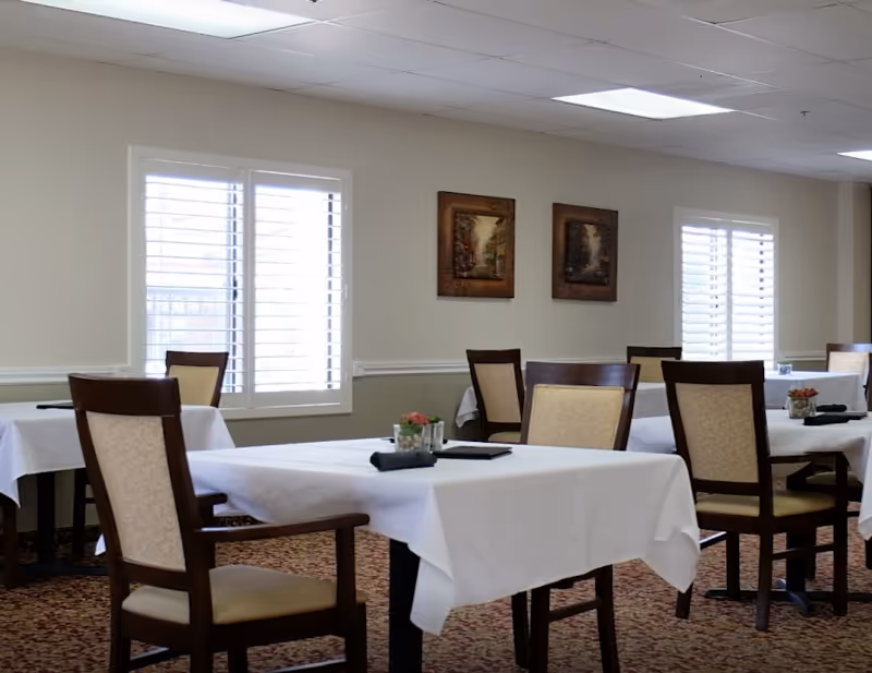 A dining room with several tables covered in white tablecloths, each set with a small flower arrangement and a folded black napkin. The room has beige walls with two framed paintings and windows with white shutters letting in natural light. The chairs have wooden frames with beige cushioned seats and backs.