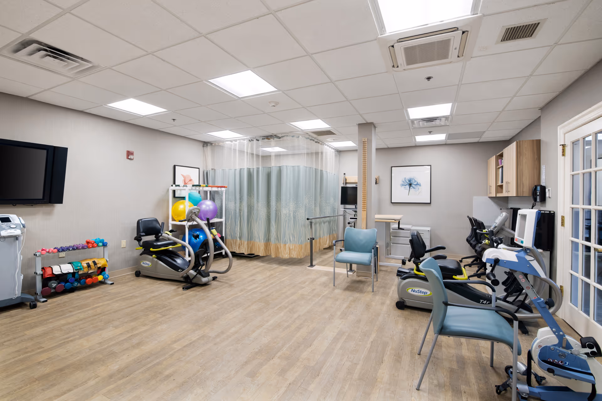 A well-lit physical therapy or rehabilitation room with exercise equipment including stationary bikes, hand weights, therapy balls, and parallel bars. The room has light wood flooring, neutral-colored walls, a mounted TV, and a privacy curtain in the back. There are chairs and cabinets along the walls, and framed artwork is displayed on the walls.