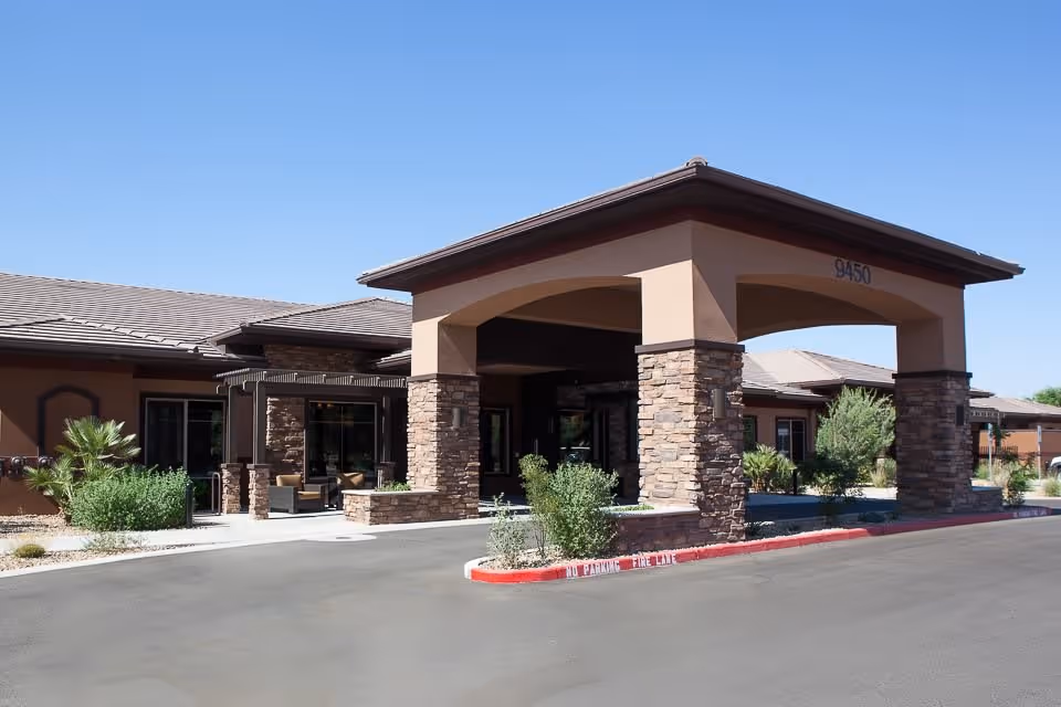 Exterior view of Mosaic Gardens at Scottsdale showing the entrance with a covered driveway supported by stone pillars, surrounded by desert landscaping and clear blue sky.