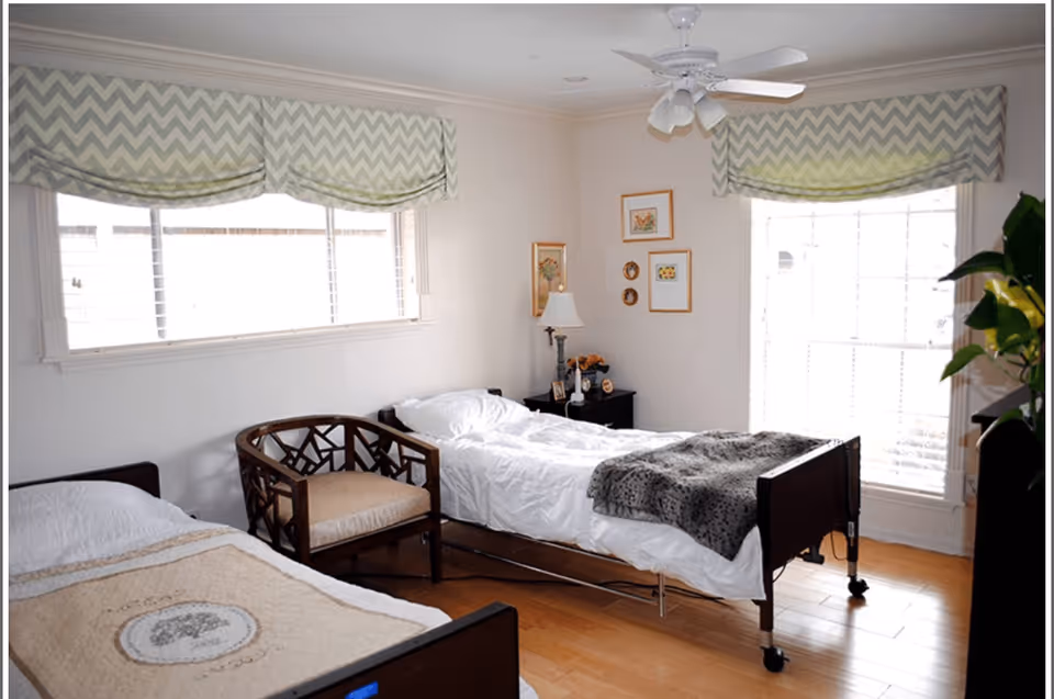 A bright bedroom with two single beds, one with a beige blanket and the other with a white blanket and a gray throw. There is a wooden chair with a cushion between the beds. The room has light-colored walls, two windows with chevron-patterned valances, a ceiling fan with lights, a small black nightstand with a lamp and decorative items, and framed pictures on the wall.