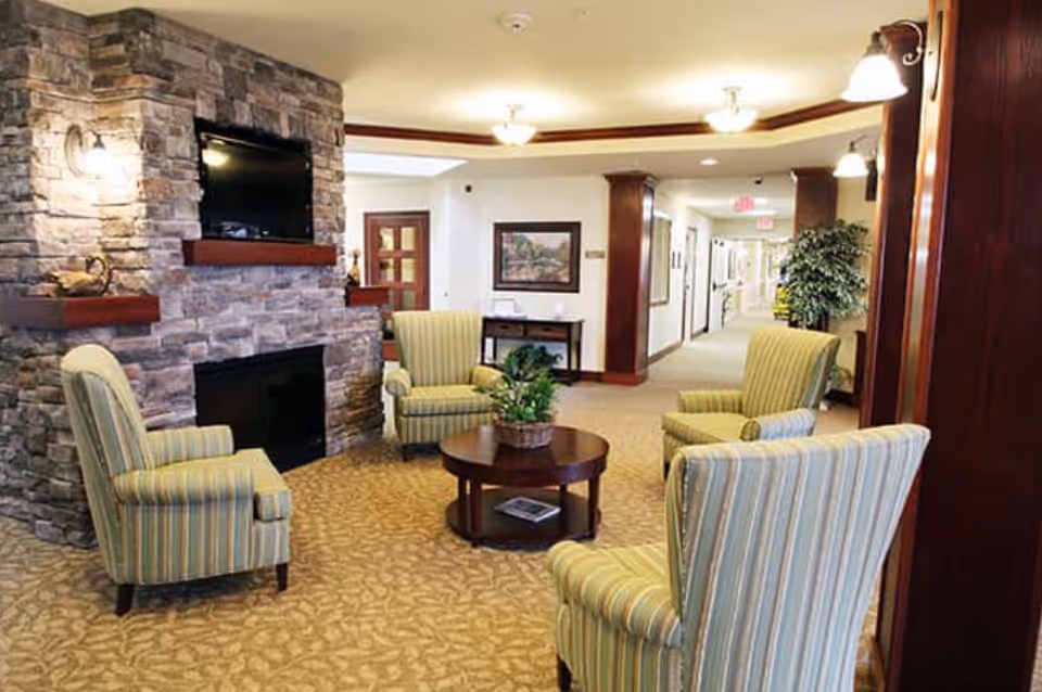 A cozy seating area in a senior living facility with four striped armchairs arranged around a round wooden coffee table with a potted plant. The room features a stone fireplace with a mounted flat-screen TV above it, warm lighting fixtures, and a carpeted floor with a patterned design. In the background, there is a hallway with framed artwork and plants.