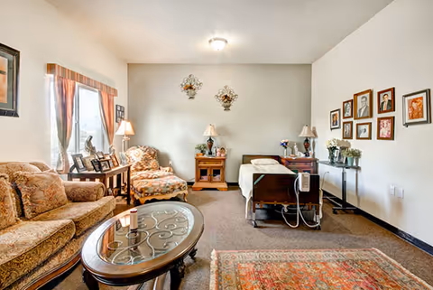 A cozy assisted living room featuring a hospital-style bed with medical equipment, a patterned armchair, a sofa, a glass coffee table, side tables with lamps, framed pictures on the walls, and decorative wall hangings. The room has a window with curtains allowing natural light to enter.