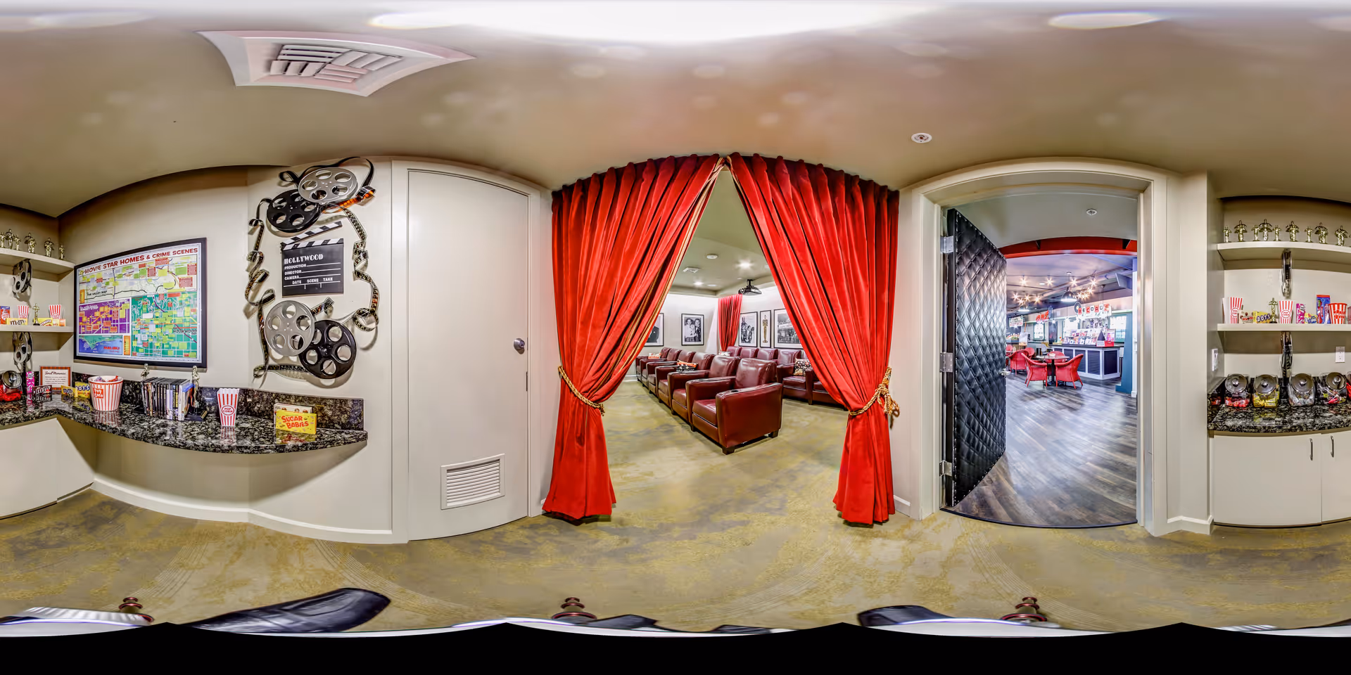 A cozy movie theater room in a senior living facility with red curtains tied back to reveal rows of brown leather recliners. To the left, a snack counter with popcorn containers, candy, and movie-themed decorations including film reels and a clapperboard. Through an open door on the right, a lounge area with red chairs and a bar counter is visible.