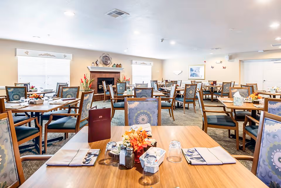 A bright and spacious dining room in a retirement community with multiple wooden tables and chairs arranged neatly. Each table is set with napkins, silverware, glasses, and small decorative centerpieces. The room features large windows with blinds, a fireplace with decorative items on the mantel, and light-colored walls with framed artwork and wall decorations.