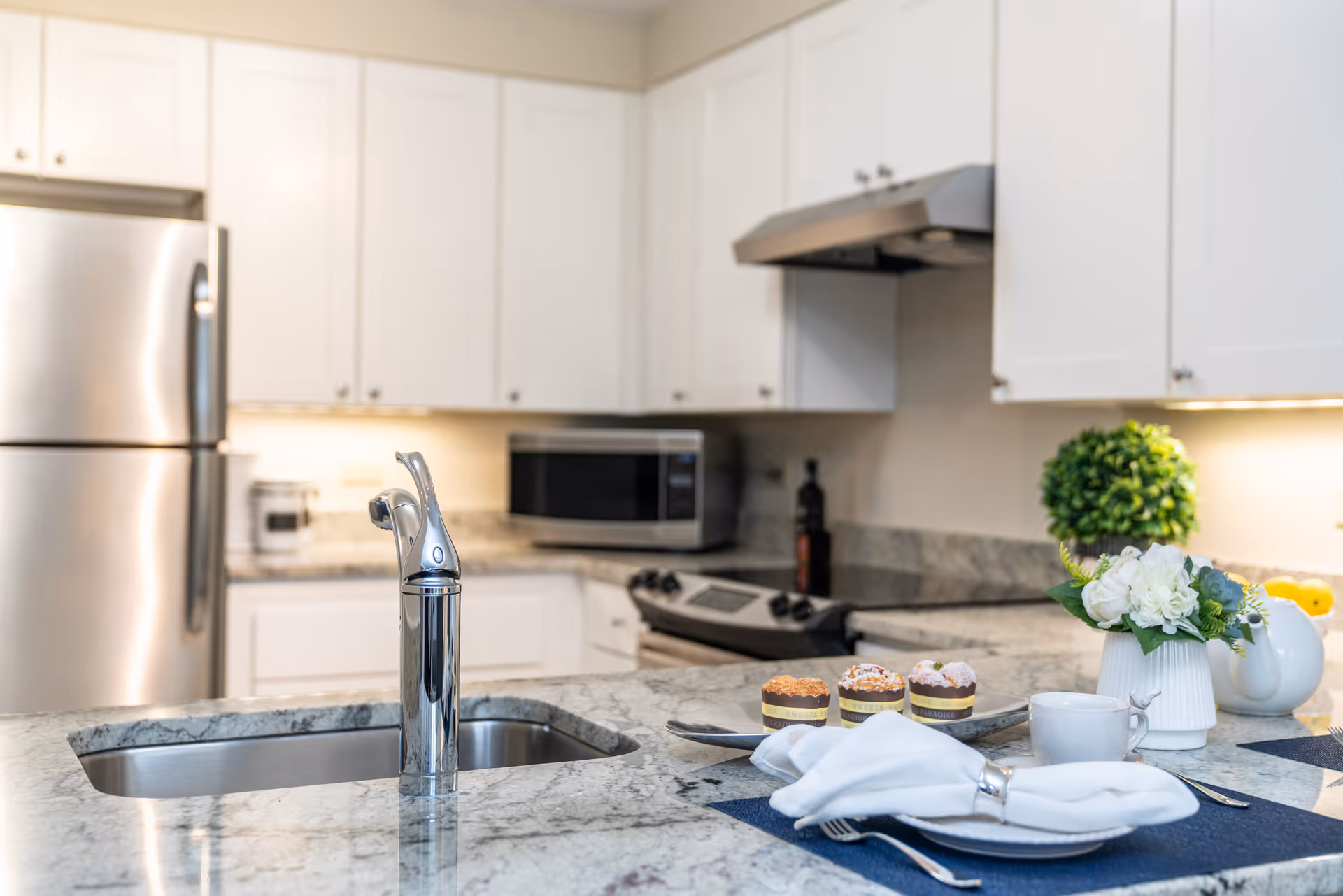 Modern kitchen with white cabinets, stainless steel refrigerator, stove, and microwave. A marble countertop island features a sink with a chrome faucet, a plate with three cupcakes, a white napkin, a cup, and a small flower arrangement.