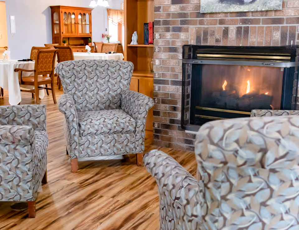 A cozy living room area with three patterned armchairs arranged around a lit brick fireplace. In the background, there is a dining area with tables covered in white tablecloths and wooden chairs, along with a wooden hutch against the wall.