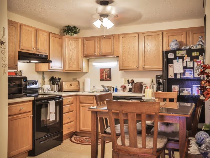 A cozy kitchen with wooden cabinets, a black stove and microwave, a black refrigerator covered with magnets and photos, a wooden dining table with four chairs, and a ceiling fan with lights. Various kitchen items and decorations are placed on the counters and table.