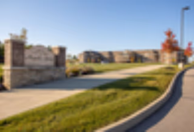 Blurred outdoor view of a senior living facility entrance with a stone sign and a paved walkway, green grass, and a building in the background under a clear blue sky.