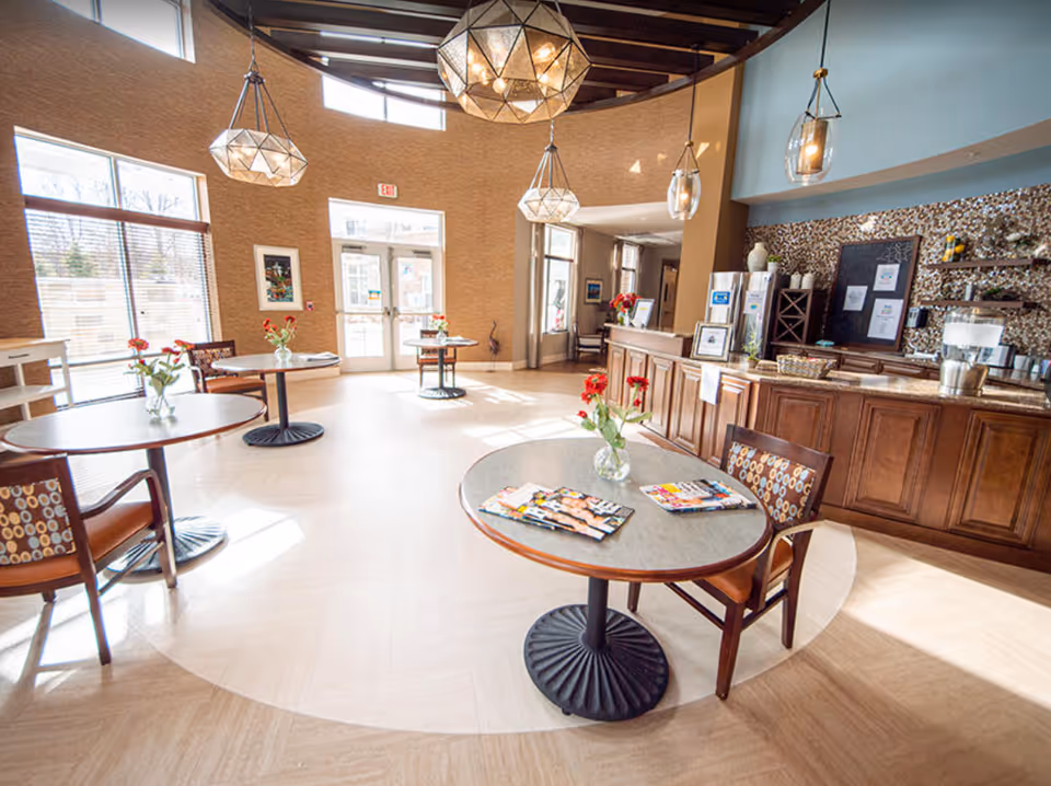 Bright communal dining area with round tables and chairs, pendant lights, and a wooden service counter.