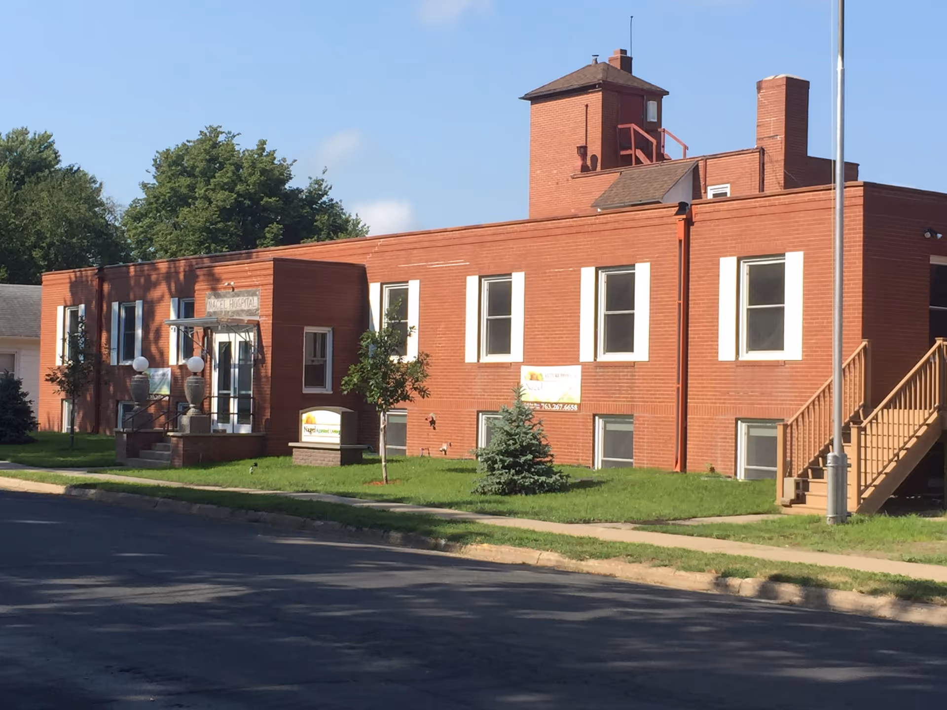 Exterior view of a red brick building with white window frames and a small entrance with stairs and two decorative lamps. The building has a sign that reads Nagel Assisted Living & Memory Care and another sign with contact information. There are small trees and grass in front of the building under a clear blue sky.