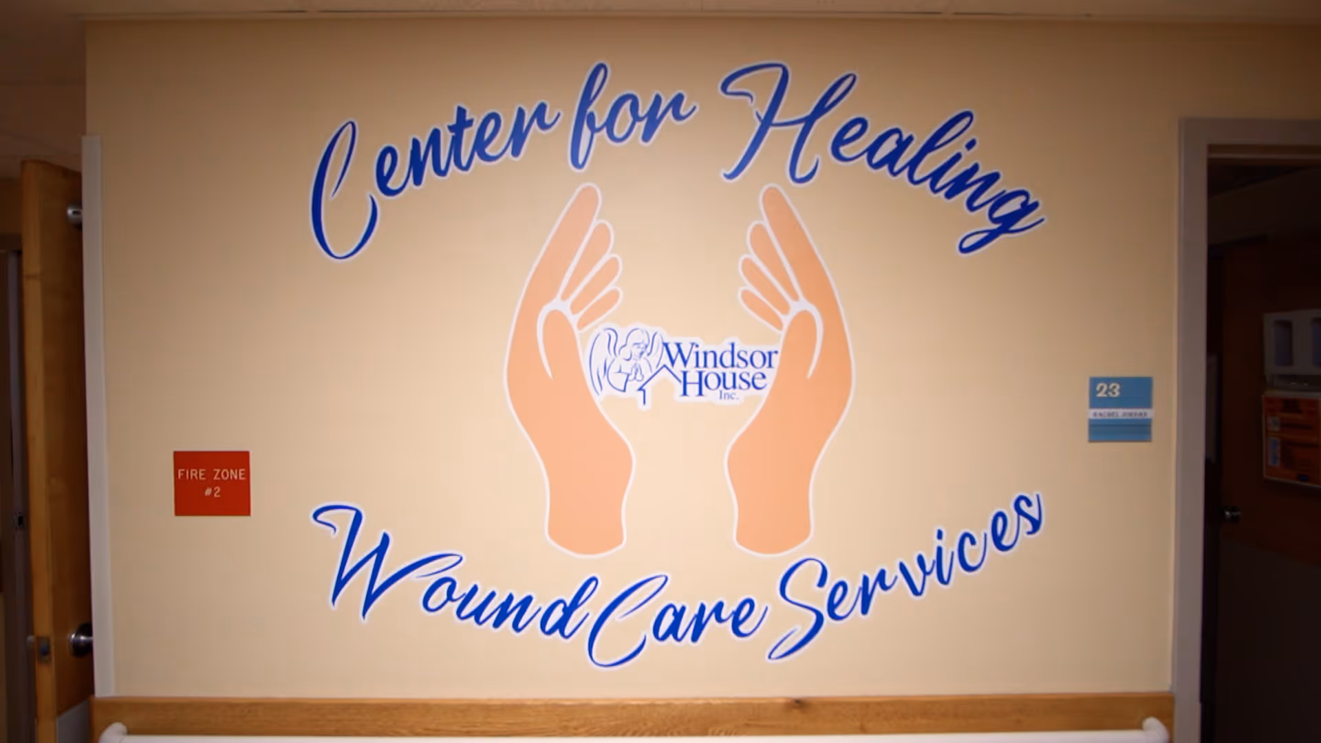 Wall mural in a healthcare facility hallway with the text 'Center for Healing Wound Care Services' surrounding an image of two hands and the Windsor House Inc. logo in the center. There is a red fire zone sign on the left and a room number sign on the right.