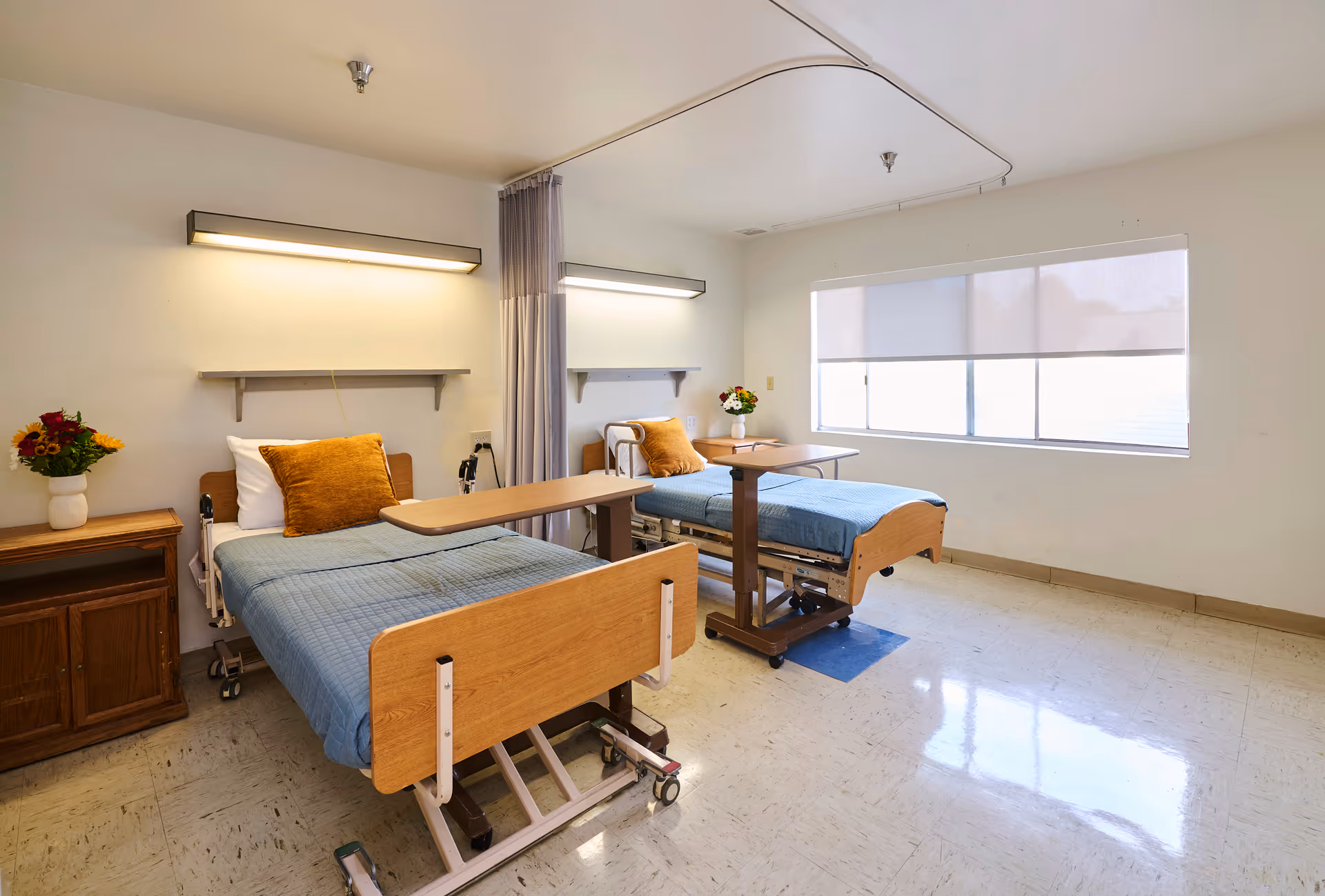 Shared senior care room with two adjustable hospital beds, overbed tables, and a large window.