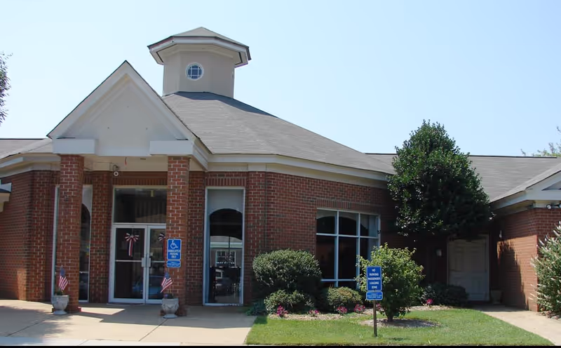 Front exterior view of a single-story brick building with a peaked roof and a small cupola on top. The entrance has double glass doors with a handicap accessible sign nearby. There are small American flags in planters on either side of the entrance and well-maintained bushes and grass in front of the building.