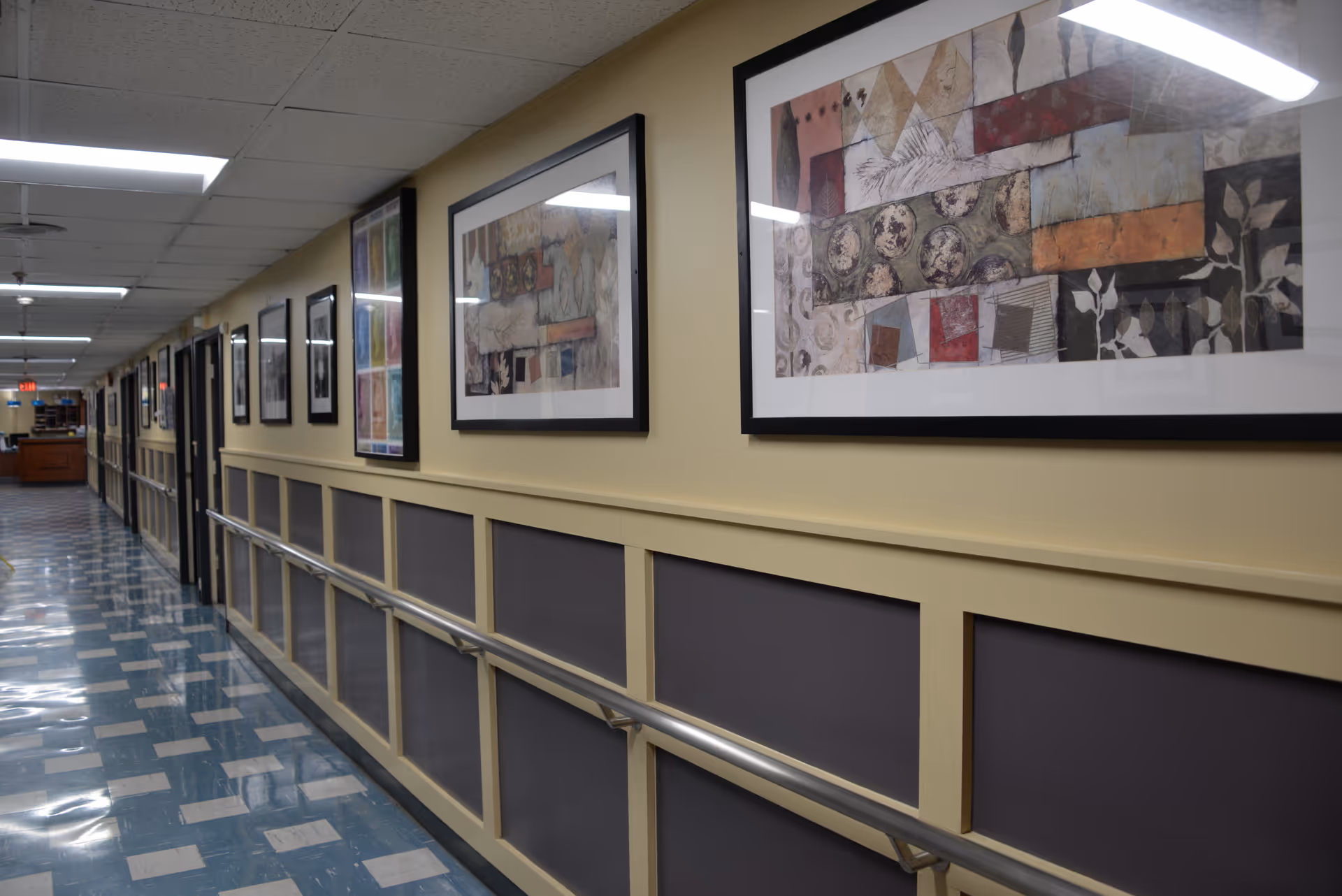 A long indoor hallway with blue and white checkered flooring, beige walls with dark panels, and handrails on both sides. The hallway is decorated with framed abstract artwork on the walls and has fluorescent ceiling lights.