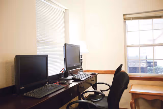 A small office or computer workstation area with two desktop computers on a dark wooden desk, two black office chairs, a table lamp, and a window with blinds partially open letting in natural light.