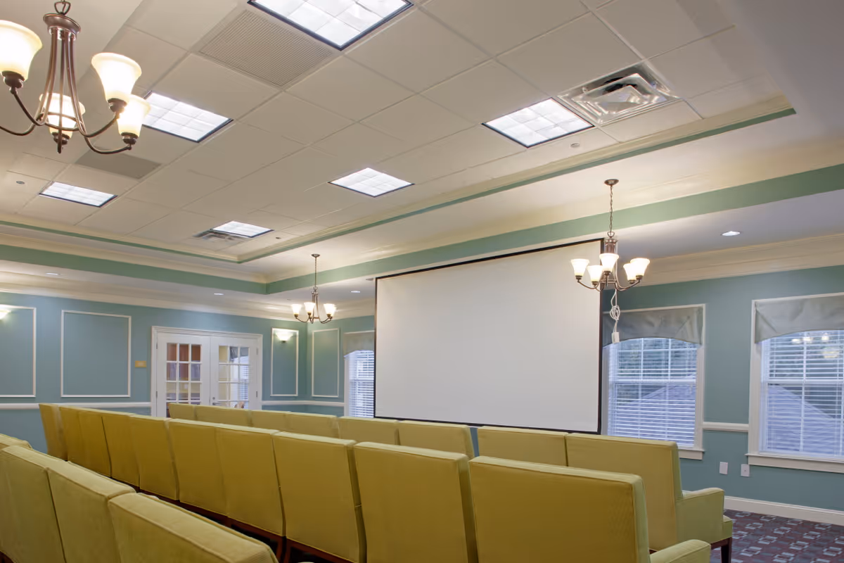 A small auditorium or meeting room with rows of green upholstered chairs facing a large white projection screen. The room has light blue walls with white trim, multiple windows with blinds, and ceiling lights including chandeliers.