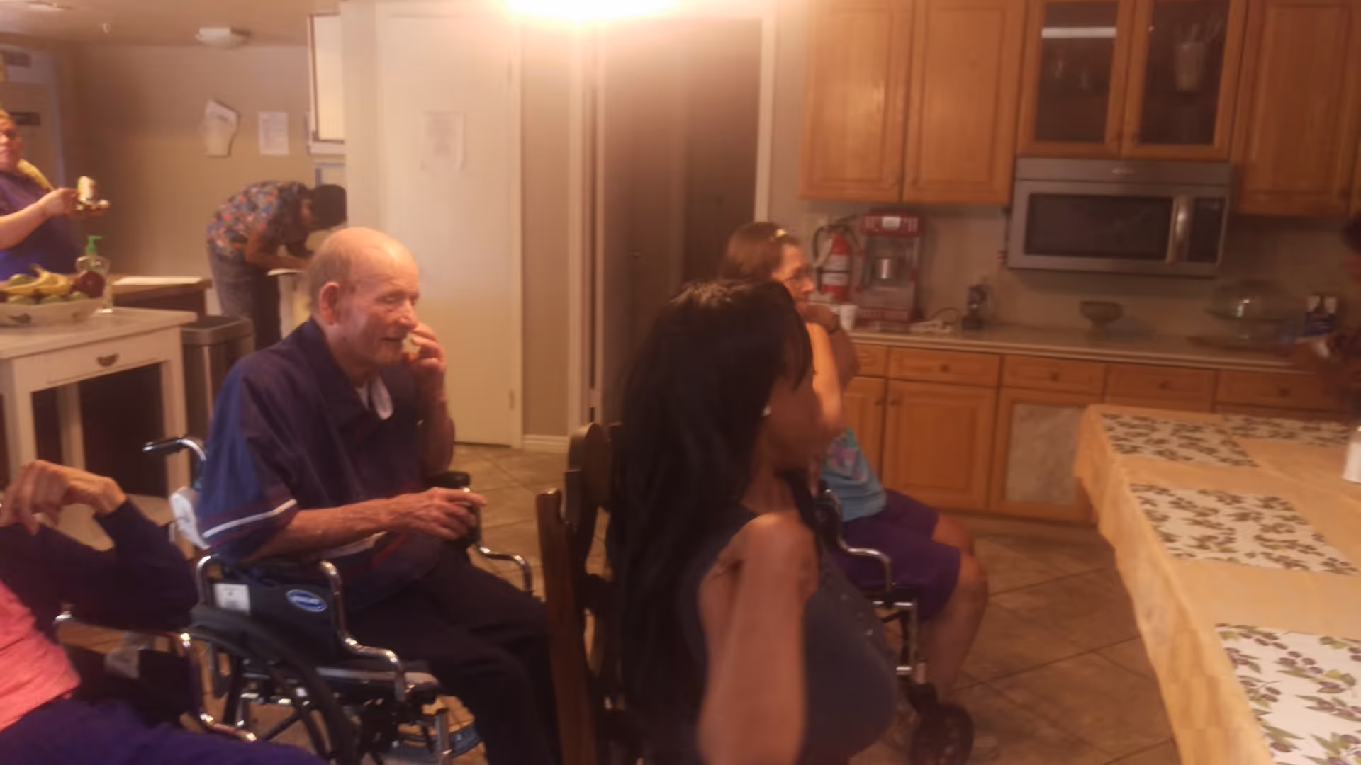 A group of elderly individuals, some in wheelchairs, gathered in a kitchen area with wooden cabinets, a microwave, and a table covered with a yellow tablecloth and placemats. One person is eating a banana in the background.