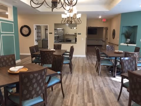 A dining room with several wooden tables and cushioned chairs arranged neatly. The room has wood flooring, light-colored walls with green accent walls, and two chandeliers hanging from the ceiling. There is a clock on one wall and a serving window or pass-through to another room.