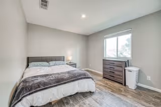 Simple bedroom with a bed, nightstand and lamp, dresser beneath a window, and wood-look flooring.