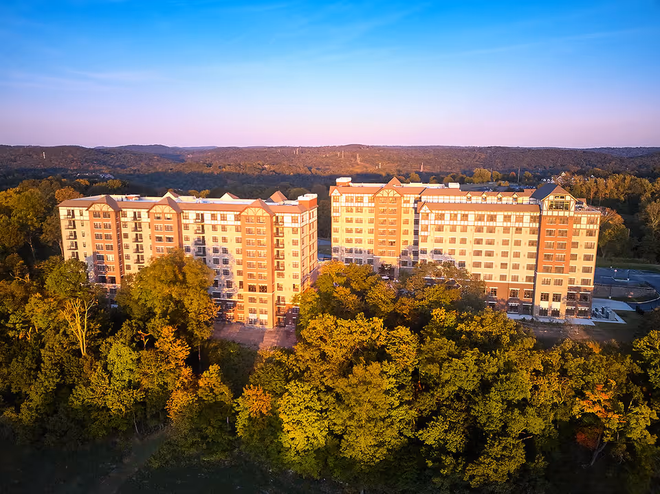 Aerial view of two large multi-story residential buildings surrounded by dense green trees with a backdrop of forested hills under a clear blue sky during sunset.