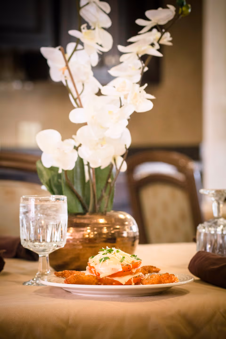 A dining table set with a plate of food featuring layered tomato and cheese garnished with herbs, a glass of water with ice, and a decorative vase with white orchids in the background.