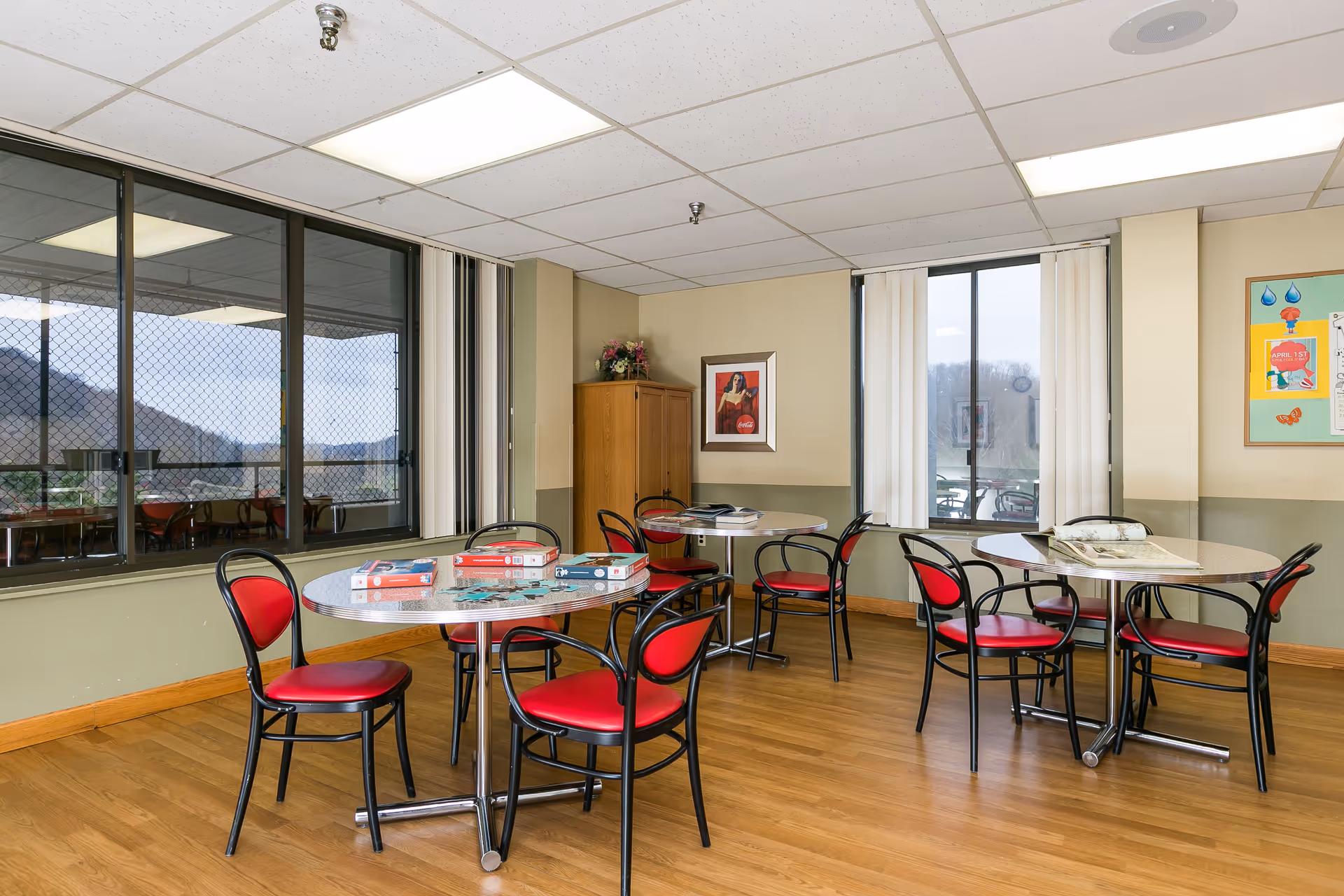 A bright room with wooden floors and beige walls featuring three round tables with black metal chairs that have red cushions. The tables have board games and magazines on them. Large windows with vertical blinds show an outdoor view with hills and a patio area. A wooden cabinet and framed pictures hang on the walls.