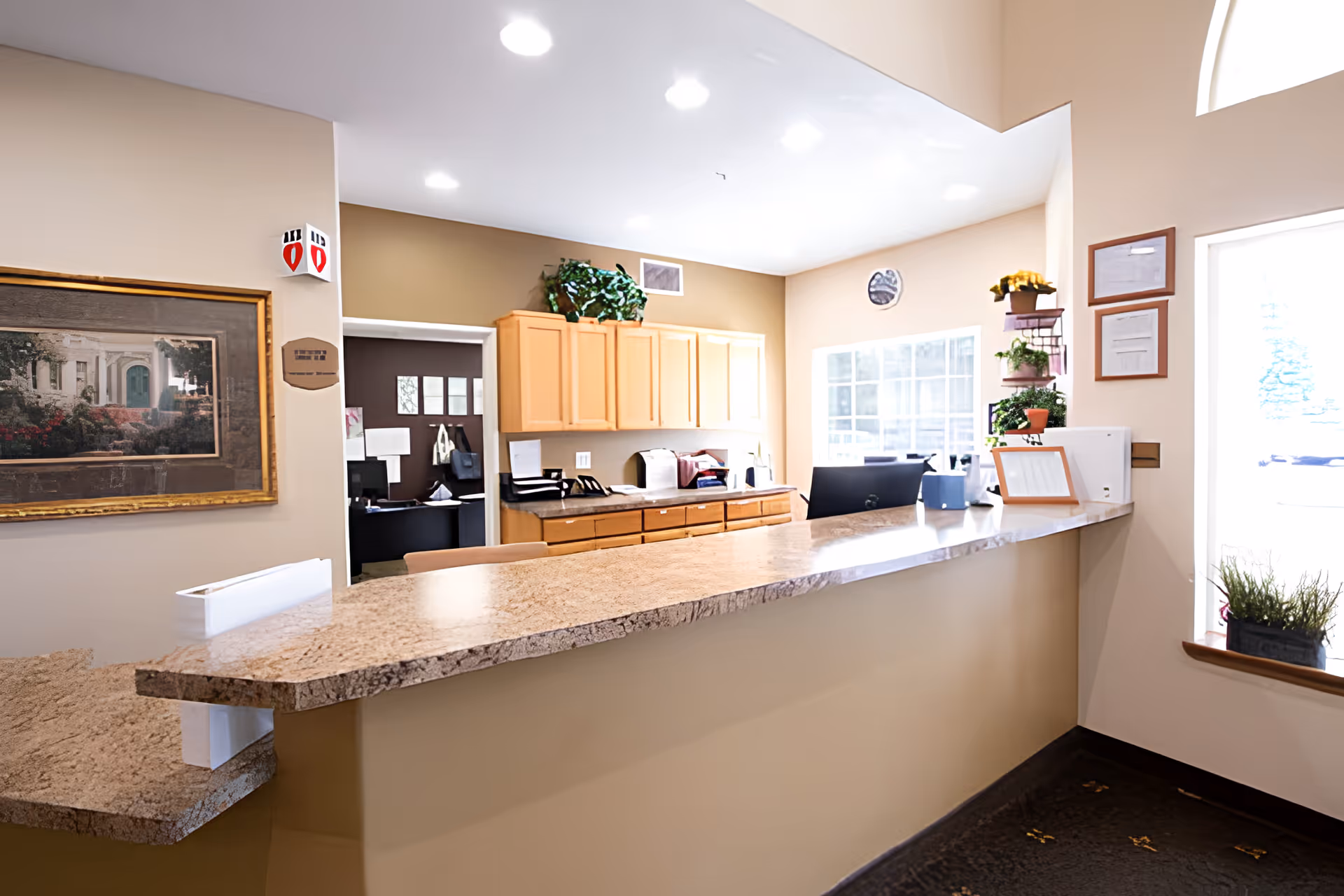 Reception area with a long granite countertop, beige walls, wooden cabinets, a computer monitor, and some potted plants near a window letting in natural light.