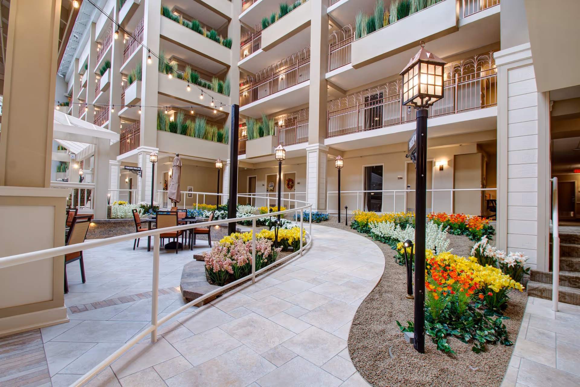 Indoor courtyard area of a senior living facility with a tiled walkway, flower beds with colorful flowers, black lamp posts, and seating areas with tables and chairs. The multi-story building has balconies with green plants and string lights hanging above.