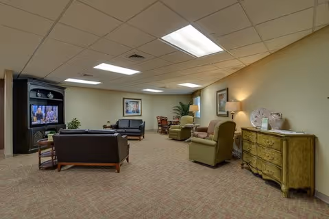 A spacious living room area with beige walls and carpeted floor. The room features several seating options including a black leather sofa, two green armchairs, and a small wooden table with chairs in the background. A large TV is placed in a black entertainment center on the left side of the room. The room is softly lit with ceiling lights and a table lamp on a green dresser on the right side. There are framed pictures and a potted plant adding decoration to the space.