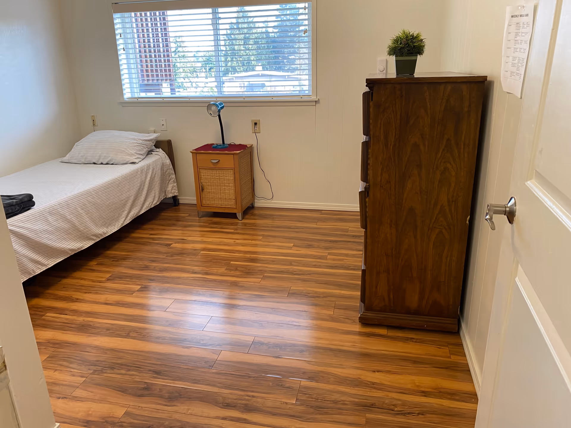 A simple bedroom with a single bed covered in a striped sheet and pillow, a small wooden nightstand with a blue lamp, a wooden dresser with a small potted plant on top, and a window with blinds letting in natural light. The floor is wooden and the walls are painted white.