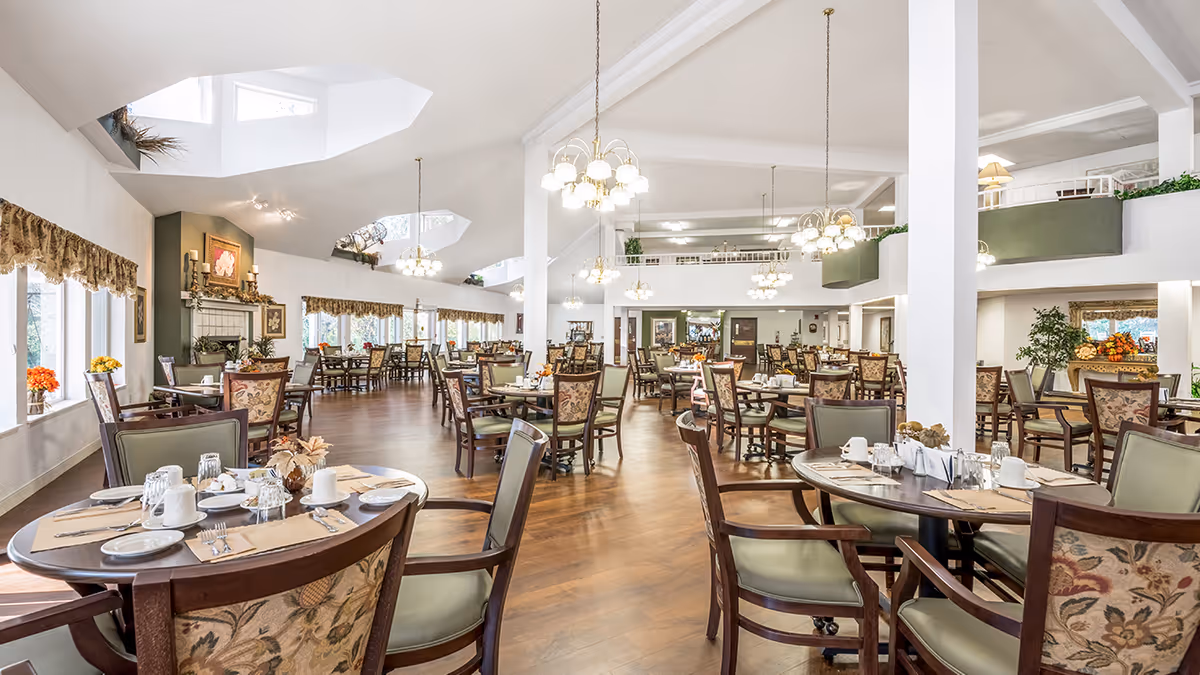 Bright, spacious dining room with multiple round tables set for meals, chandeliers, high ceilings, and wood flooring.