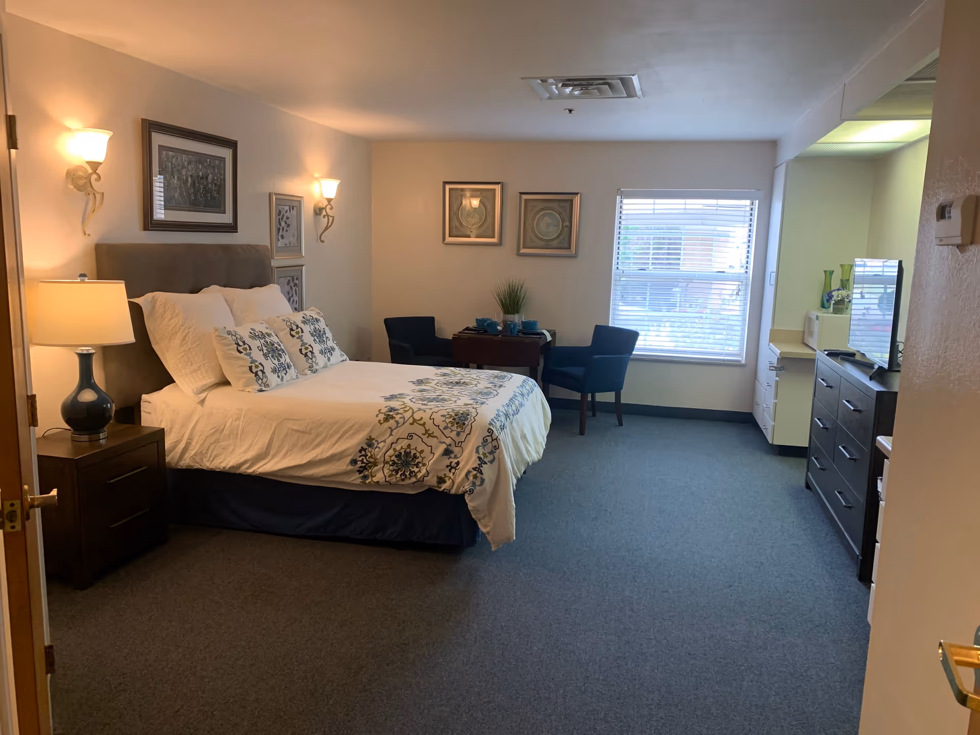 A cozy bedroom in an assisted living facility featuring a neatly made bed with white and blue patterned bedding, two wall-mounted lamps above the bed, a bedside table with a lamp, two blue chairs with a small table between them near a window with blinds, a dresser with a TV on top, and a small kitchenette area with a microwave and decorative vases.