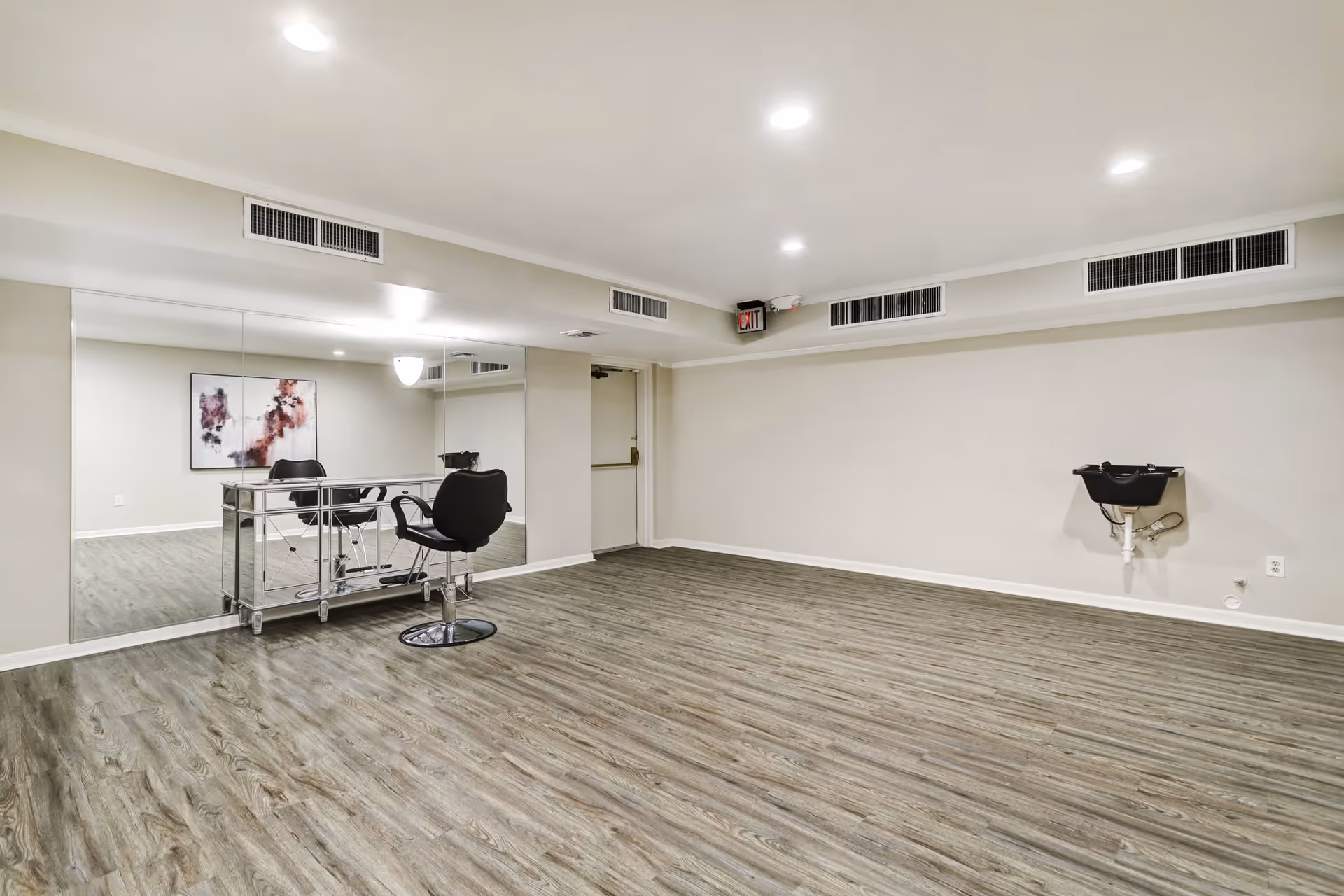 A spacious room with wood-patterned flooring and light-colored walls. There is a large mirror on one wall reflecting a black salon chair and a mirrored styling station. On the opposite wall, there is a black wash basin mounted. The ceiling has recessed lighting and air vents, and an exit sign is visible above a door.