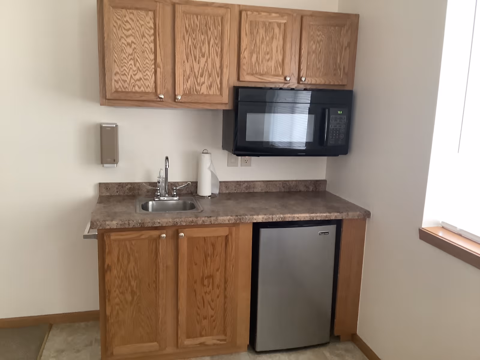 Small kitchenette with wooden cabinets, a countertop sink, an over-the-range microwave, and a mini refrigerator.
