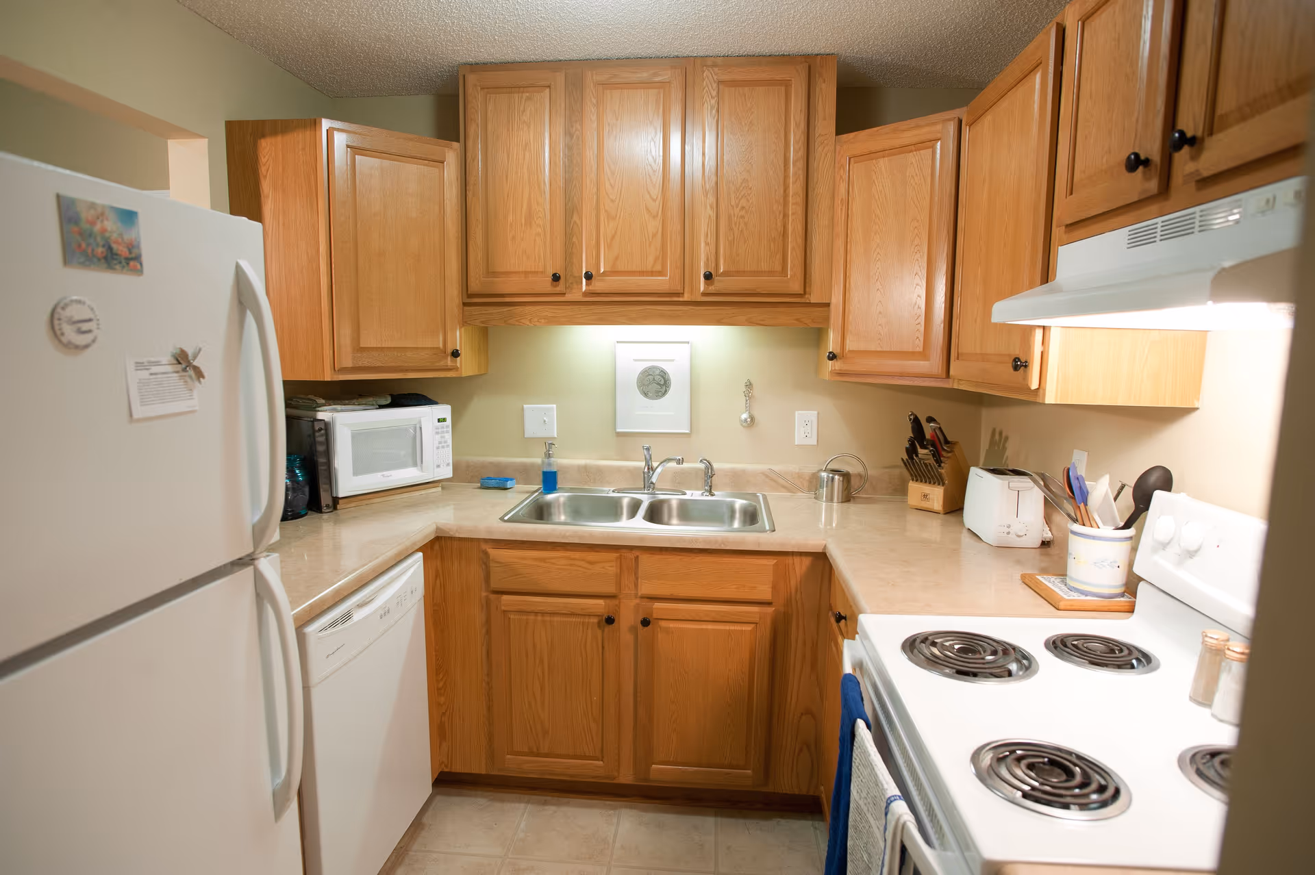 A compact kitchen with wooden cabinets, a white refrigerator, a microwave, a dishwasher, a double sink, a white electric stove with coil burners, a toaster, a knife block, and various kitchen utensils on the countertop.