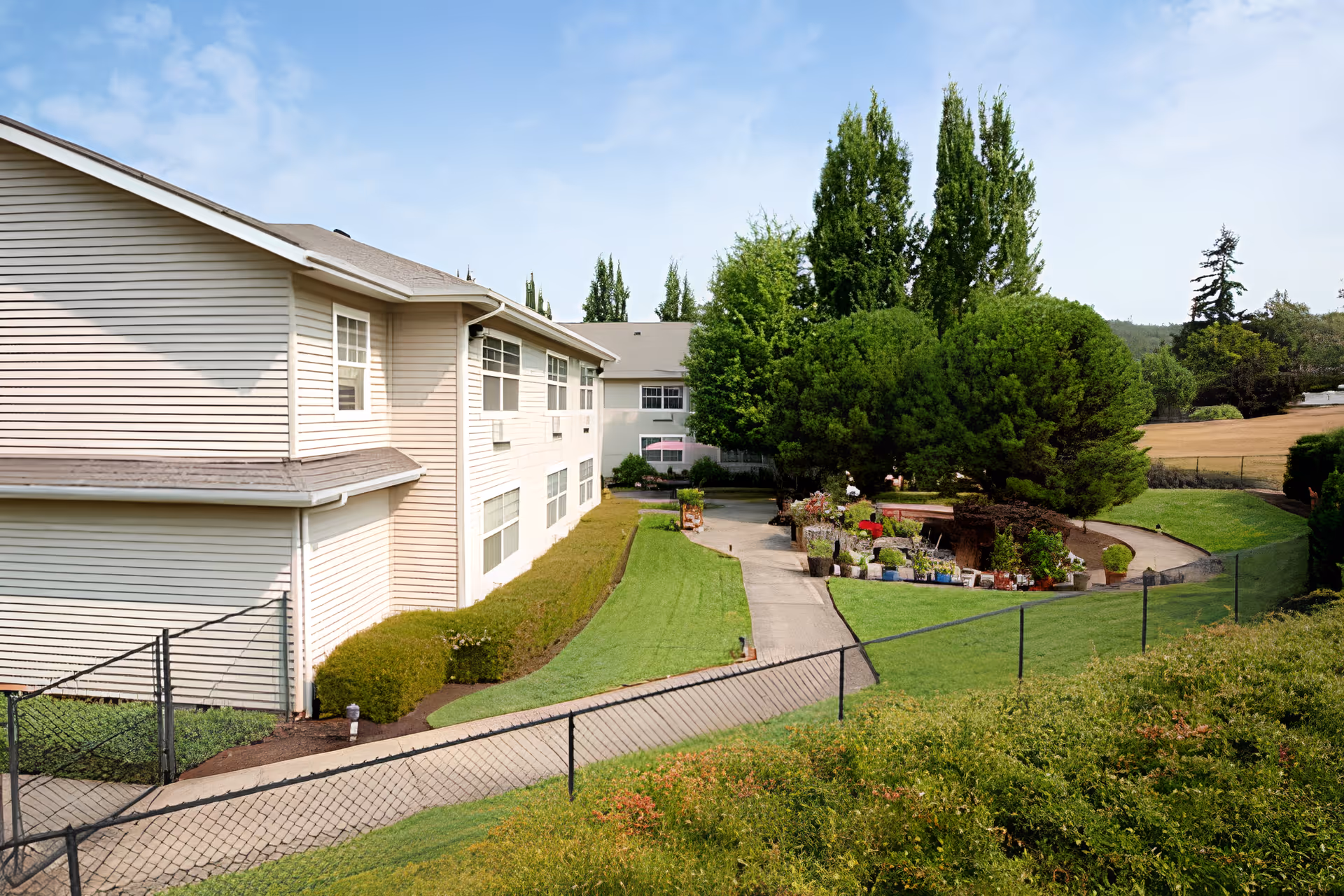 Exterior view of a senior living facility building with beige siding, surrounded by well-maintained green lawns, bushes, and tall trees under a clear blue sky. A paved pathway runs alongside the building and leads to a garden area with various potted plants and shrubs.