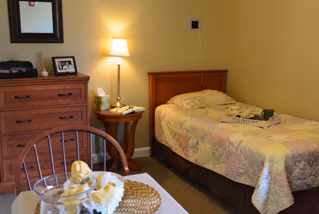 A cozy bedroom in a senior living facility with a single bed covered in a pastel patchwork quilt. Next to the bed is a wooden nightstand with a lamp, tissue box, and a book. A wooden dresser with a framed photo and decorative items is against the wall. In the foreground, part of a dining table with a woven placemat and a glass pitcher is visible.