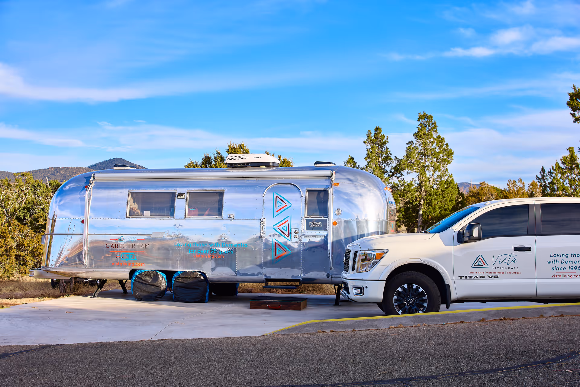 A shiny silver trailer parked on a concrete pad next to a white pickup truck with Vista Living Care branding. The trailer has windows and colorful triangular designs on its side. Trees and hills are visible in the background under a blue sky.