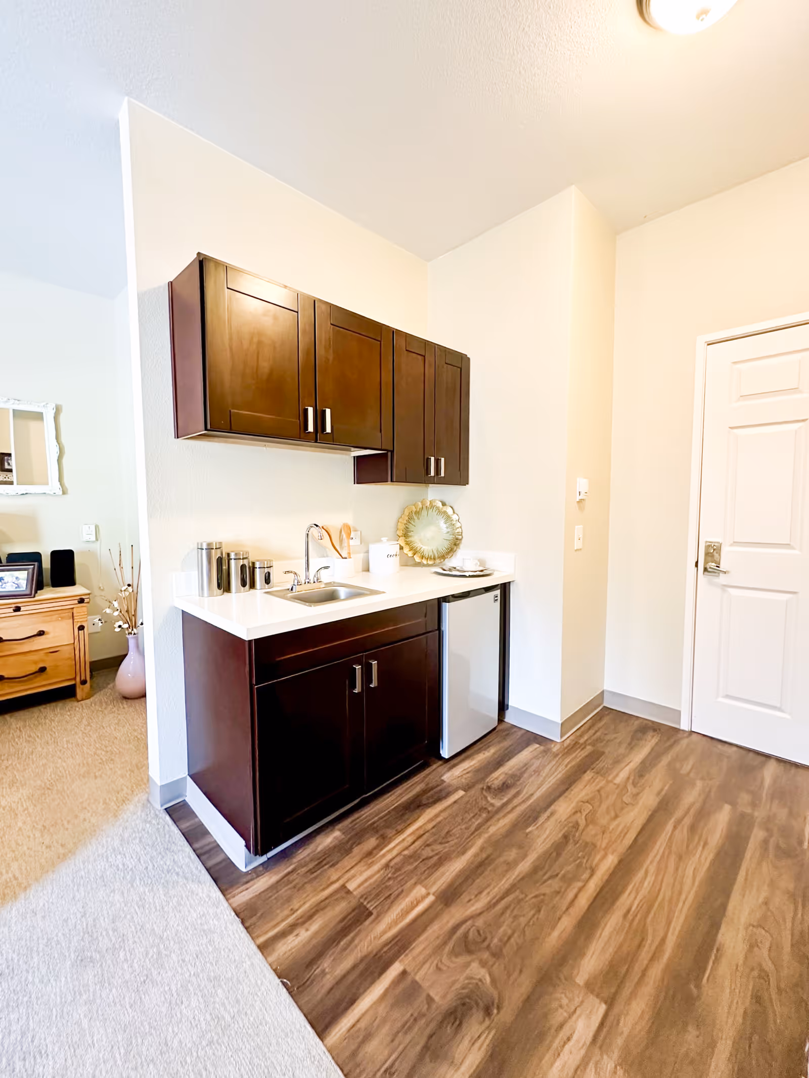A small kitchenette area with dark wooden cabinets, a white countertop, a stainless steel sink, and a mini refrigerator. The floor is wood laminate, and there is a white door to the right. Part of a living area with a wooden dresser and a mirror is visible to the left.