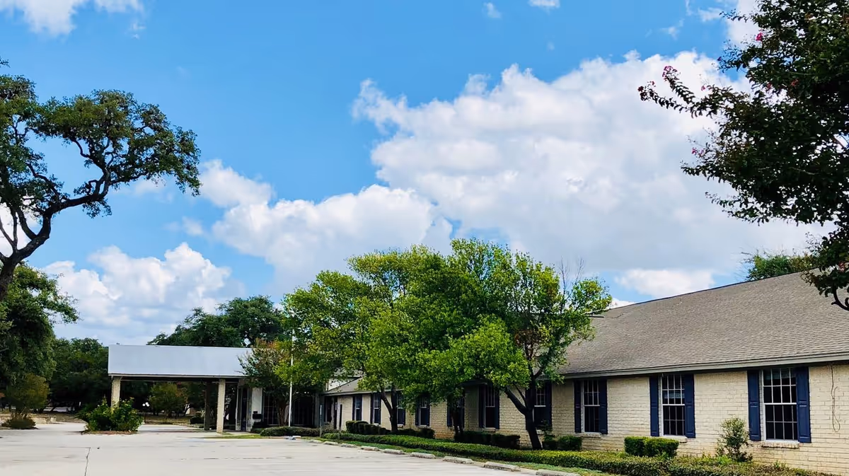 Exterior view of a single-story building with a light-colored brick facade and blue window shutters, surrounded by green trees under a partly cloudy blue sky. There is a covered entrance area and a paved driveway in front.