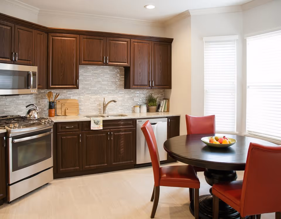 A modern kitchen with dark wooden cabinets, a stainless steel oven and microwave, a sink with a faucet, and a dishwasher. There is a round dark wooden dining table with four red chairs around it, and a bowl of fruit on the table. The room is well-lit with natural light coming through two large windows with white blinds.