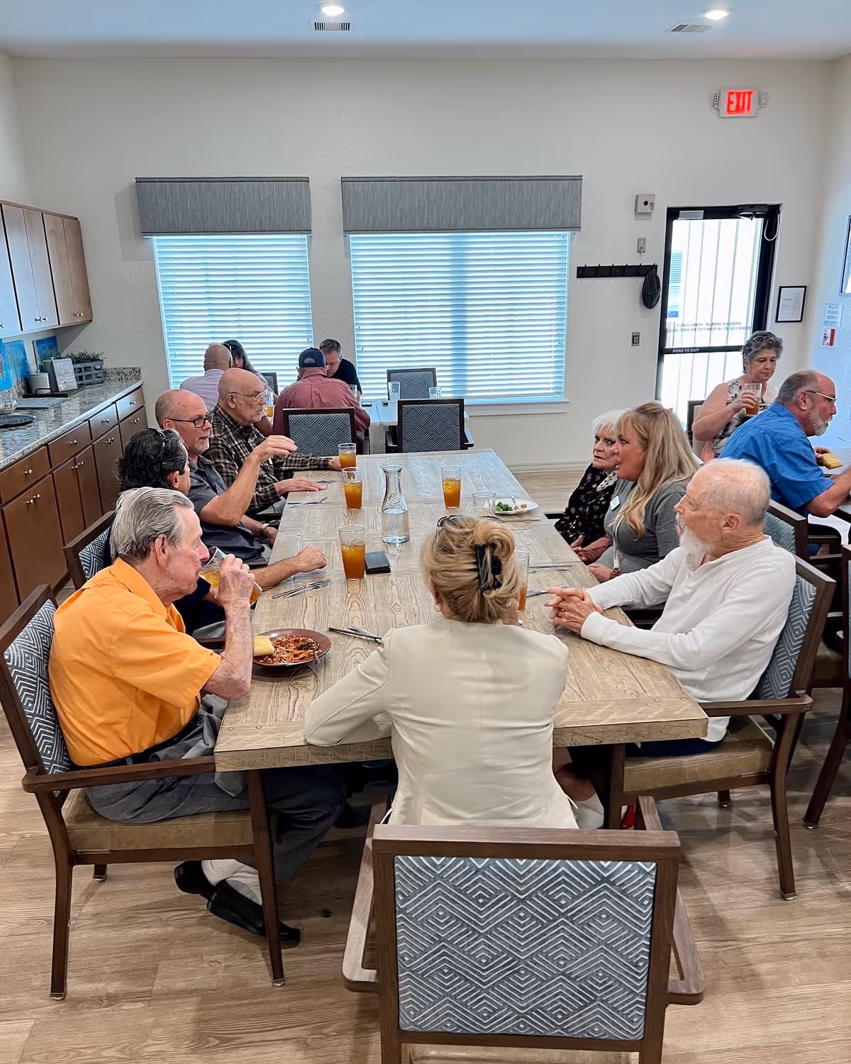 A group of elderly people sitting around a large wooden dining table in a bright room with windows covered by blinds. They are drinking iced tea and eating food, engaged in conversation. The room has wooden cabinets along one wall and a door with an exit sign above it.