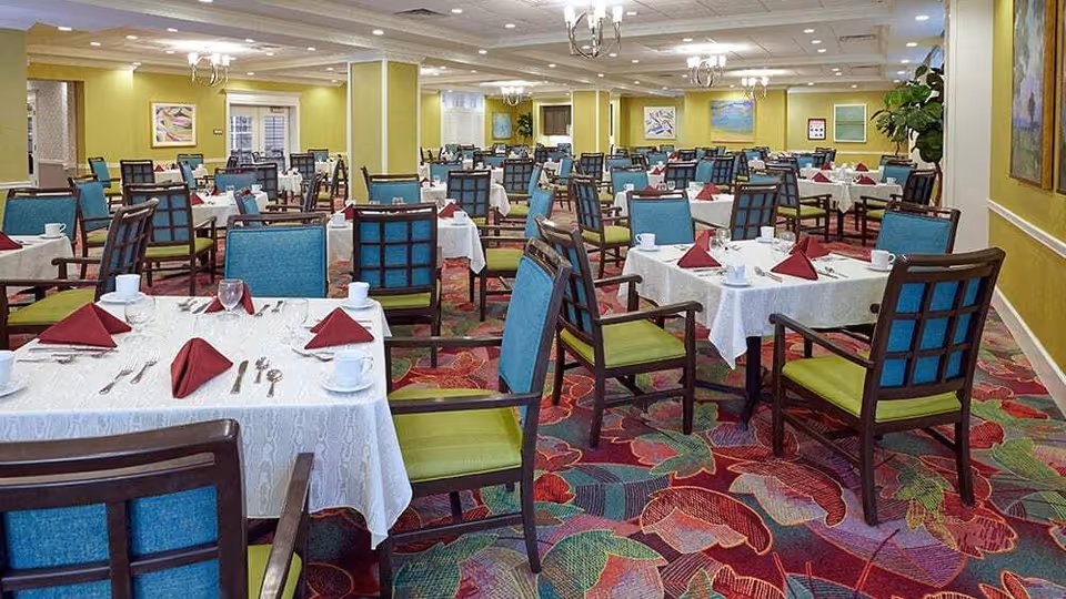 A spacious dining room with multiple tables covered in white tablecloths, each set with red folded napkins, cups, glasses, and silverware. The chairs have blue backs and green seats. The room has yellow walls adorned with framed artwork and a colorful carpet with a floral pattern.