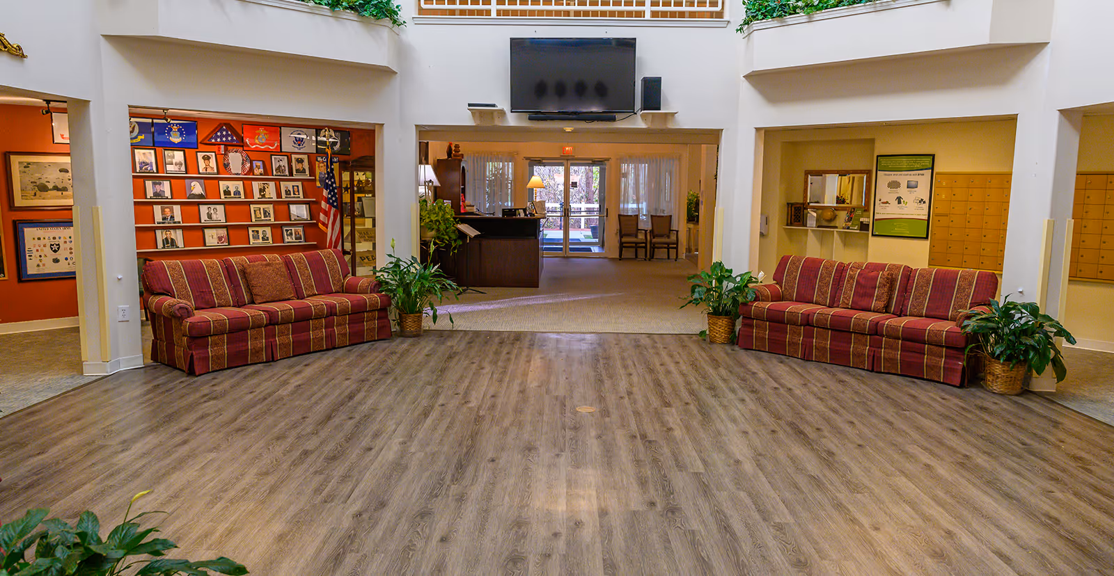 Spacious senior living facility lobby with two striped sofas, potted plants, a reception desk, and a wall-mounted TV.