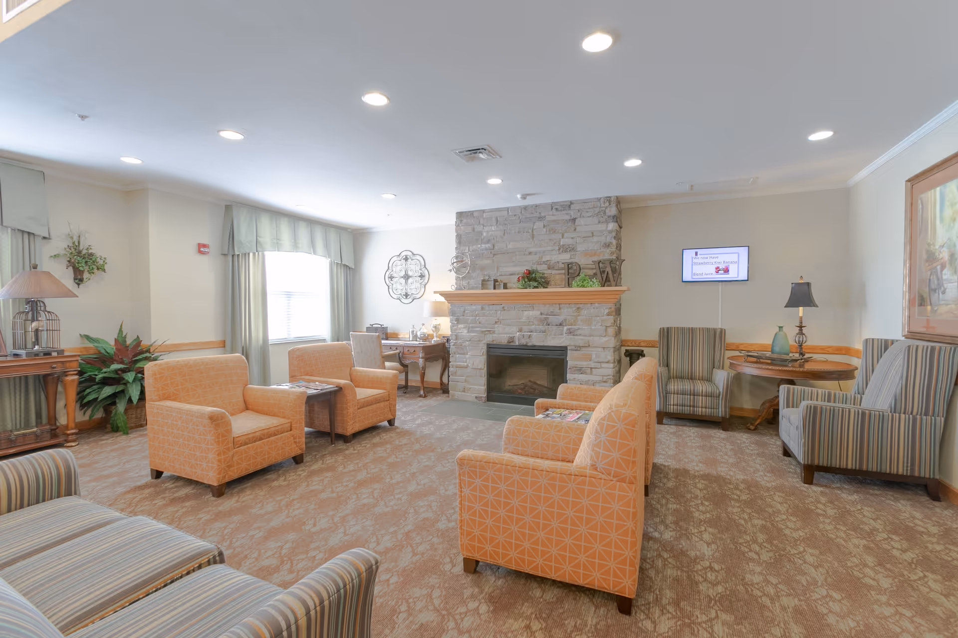 A cozy living room area with multiple armchairs in orange and striped patterns arranged around a stone fireplace. The room has beige walls, a window with green curtains, a small wooden table with a lamp, and decorative plants. A flat-screen TV is mounted on the wall displaying a message about juice flavors.