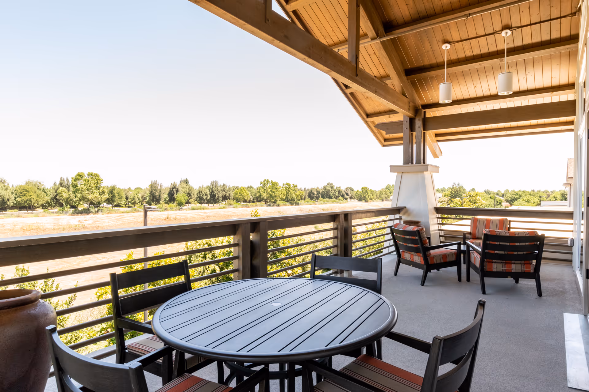 Covered outdoor patio area with a round table and four chairs in the foreground, and a seating area with four cushioned chairs and a small table in the background, overlooking a landscape of trees and open land under a clear sky.