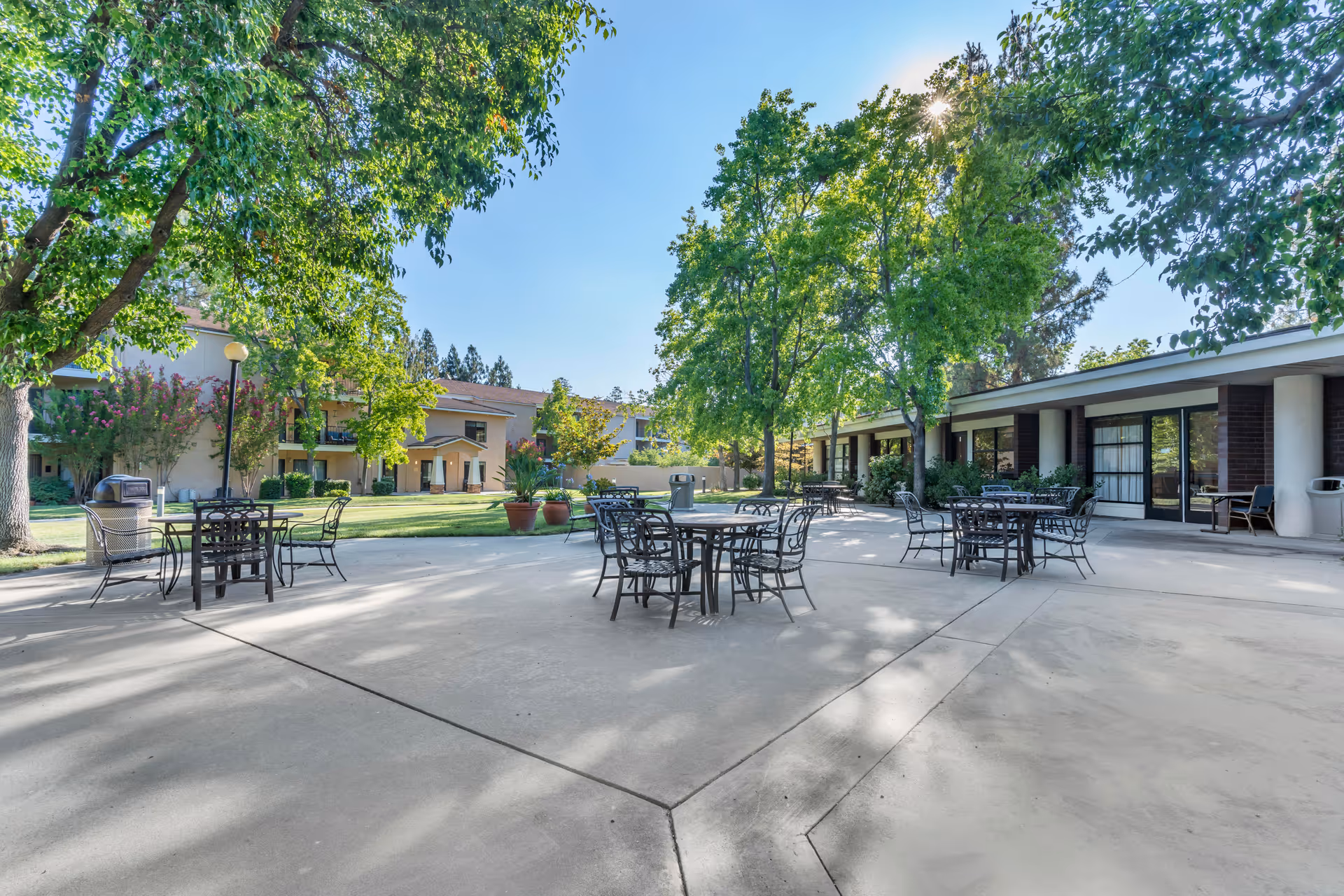 Outdoor patio area at Brookdale San Jose with several metal tables and chairs arranged on a concrete surface. The patio is surrounded by green trees and plants, with a two-story building visible in the background under a clear blue sky.