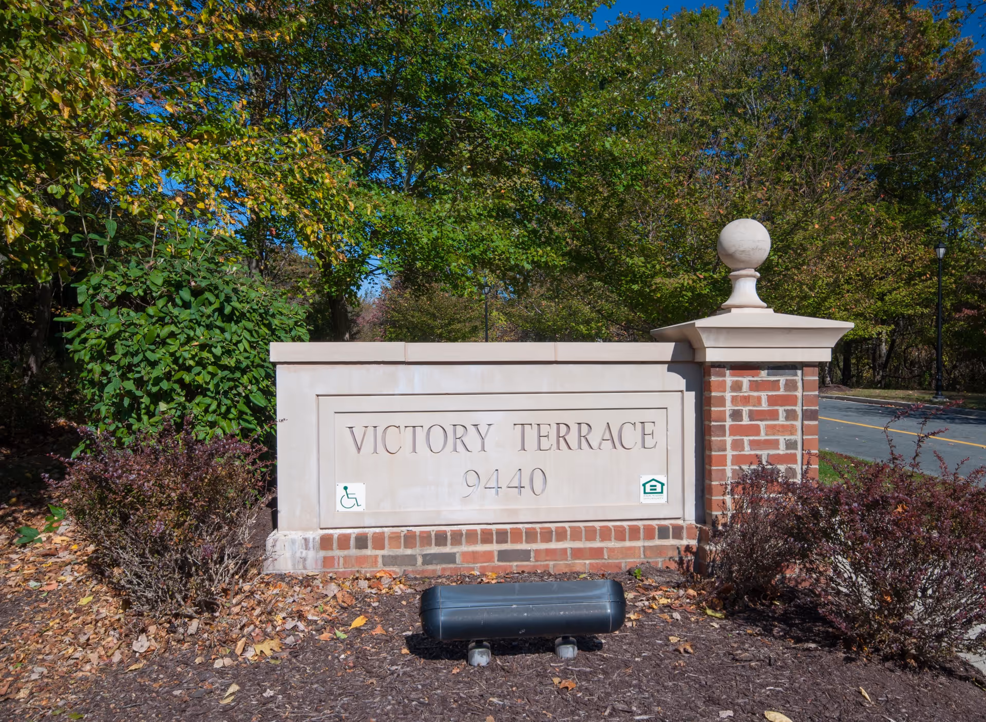 Stone and brick sign for Victory Terrace with the number 9440, surrounded by bushes and trees with green leaves under a clear blue sky.