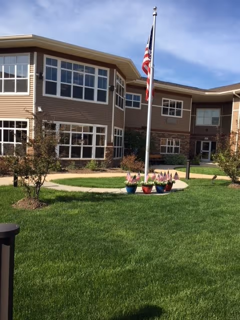 Exterior view of a two-story senior living facility building with large windows, beige siding, and stone accents. In front of the building is a well-maintained green lawn with a circular concrete path surrounding a flagpole flying the American flag. Several small potted plants with miniature American flags are placed around the base of the flagpole. The sky is clear with some clouds.