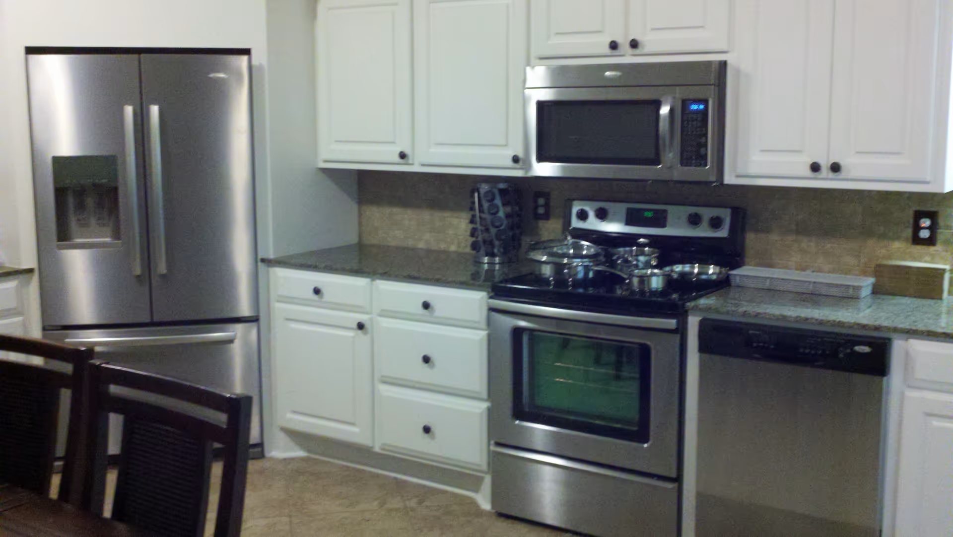 Modern kitchen with stainless steel appliances including a refrigerator, microwave, stove, and dishwasher. White cabinets with black knobs and granite countertops are visible. A spice rack and some cookware are on the stove. Part of a dining table with chairs is seen in the foreground.